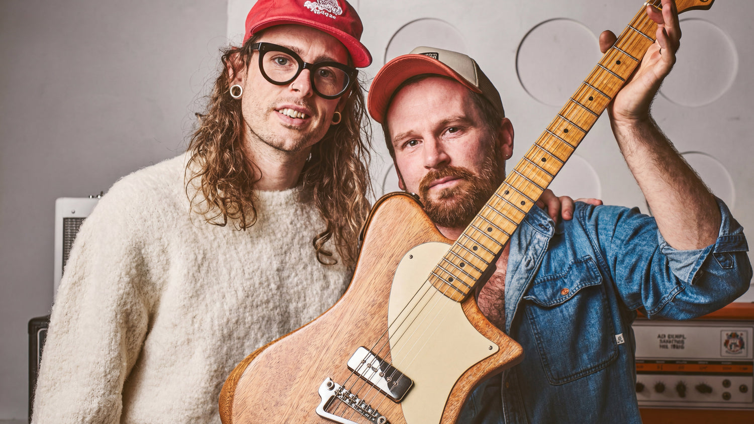 Two men, one holding a guitar, standing in front of audio equipment.